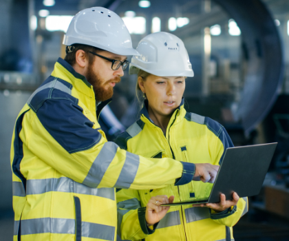 A man and woman, both in high-visibility jackets and hard hats, looking at a laptop together in an industrial setting